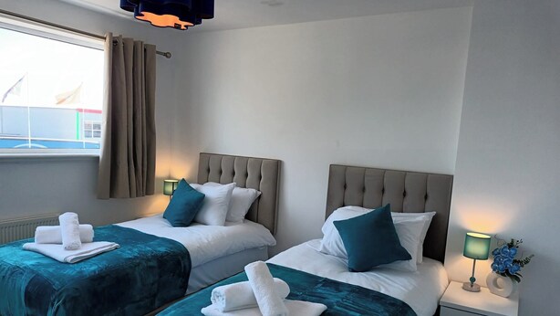 A hotel room with two beds, each with a teal bedspread and white pillows, and a nightstand with a lamp and a vase of flowers. - Charming 3-bed House in Birmingham Near nec & hs2 (Birmingham)