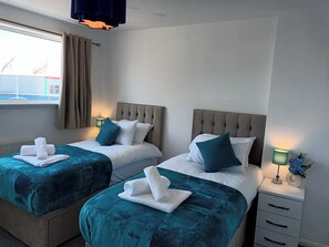 A hotel room with two beds, each with a teal bedspread and white pillows, and a nightstand with a lamp and a vase of flowers. - Charming 3-bed House in Birmingham Near nec & hs2 (Birmingham)