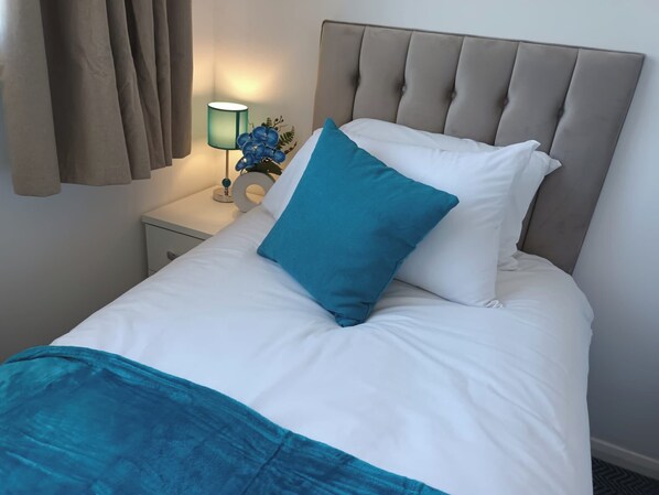 A neatly made bed with white and teal bedding, a grey upholstered headboard, and a bedside lamp with a blue flower decoration. - Charming 3-bed House in Birmingham Near nec & hs2 (Birmingham)