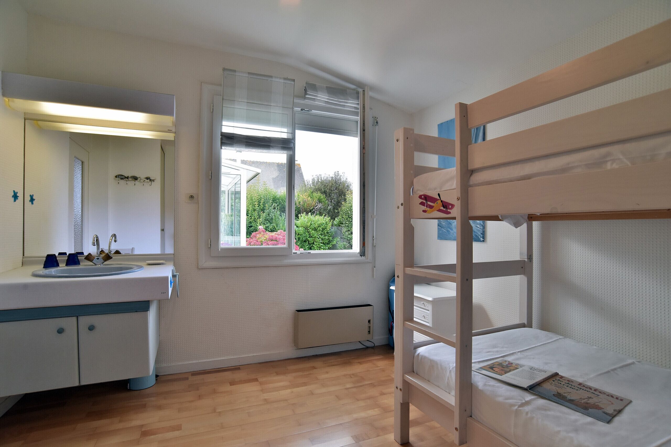 Een slaapkamer met een stapelbed, een raam met uitzicht op groen, een witte wastafel met blauwe accenten en een spiegel.