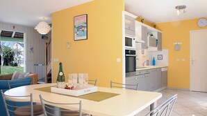 Un intérieur lumineux aux murs jaune vif, avec une table à manger dressée pour quatre personnes, une cuisine aux meubles blancs et une grande fenêtre donnant sur un jardin.