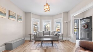 A living room with a grey sofa, a coffee table, and a bench. There are three windows, a door with a wreath, and a chandelier. - Luxe Ohio City Townhome only minutes to stadium! (Cleveland)