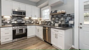A modern kitchen with white cabinets, a stainless steel microwave, oven, and dishwasher, a tiled backsplash, and a window with blinds. - Luxe Ohio City Townhome only minutes to stadium! (Cleveland)