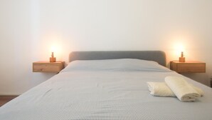 Una cama bien hecha con una colcha a rayas y una toalla enrollada, flanqueada por dos mesitas de noche con lámparas, frente a una pared lisa.