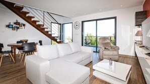 Un salon moderne avec un canapé d’angle, un coin repas doté d’une table ronde, un escalier et de grandes fenêtres offrant une vue sur l’extérieur.