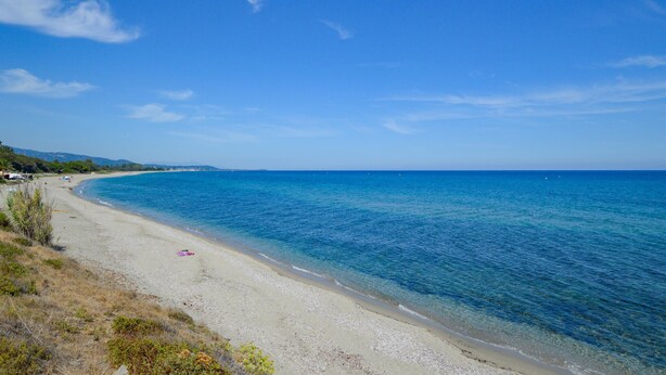 Une plage avec une eau d’un bleu limpide, un rivage sablonneux et quelques personnes visibles au loin.