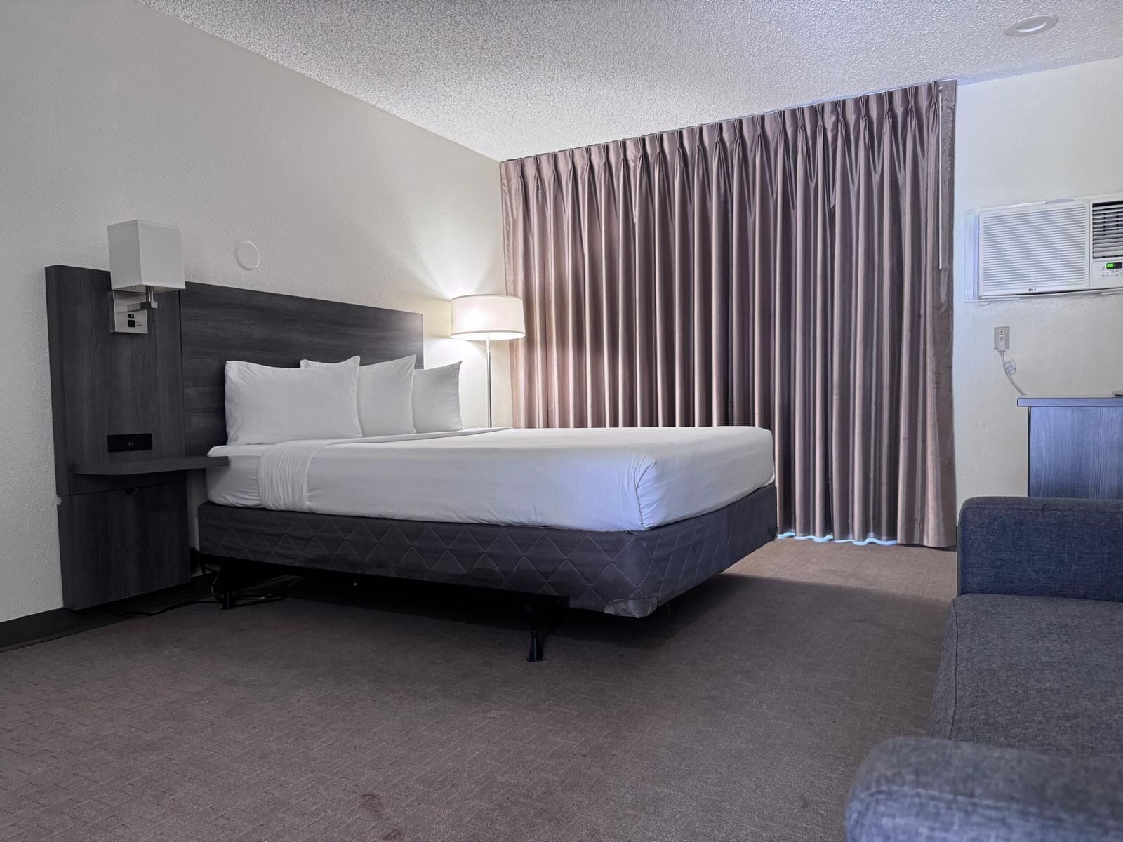 Une chambre d’hôtel comprenant un lit, une table de chevet, une lampe, un téléviseur, une fenêtre avec des rideaux et un climatiseur fixé au mur.
