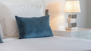 Ein ordentlich bezogenes Bett mit weißen und blauen Kissen, einer Nachttischlampe und einer kleinen Uhr auf dem Nachttisch.