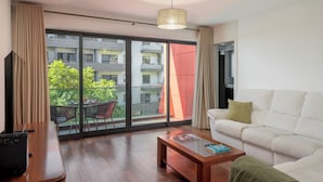 Ein modernes Wohnzimmer mit einer Couch, einem Couchtisch, einem Fernseher und einem großen Fenster mit Blick auf einen Balkon und Gebäude.