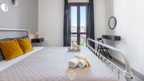 A neatly made bed with rolled towels, a bedside table with a lamp, and a view of the outside through a window with curtains.
