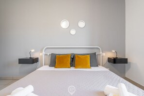 Quarto moderno com cama branca, duas mesas de cabeceira e luminária fixada na parede.