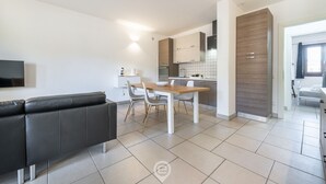 A modern living room with a black leather sofa, a wooden dining table with chairs, and a kitchen area with white cabinets and a tiled backsplash.