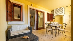 A cozy outdoor seating area with a sofa and dining table, a sliding glass door leading to a bathroom, and an air conditioning unit on the wall.