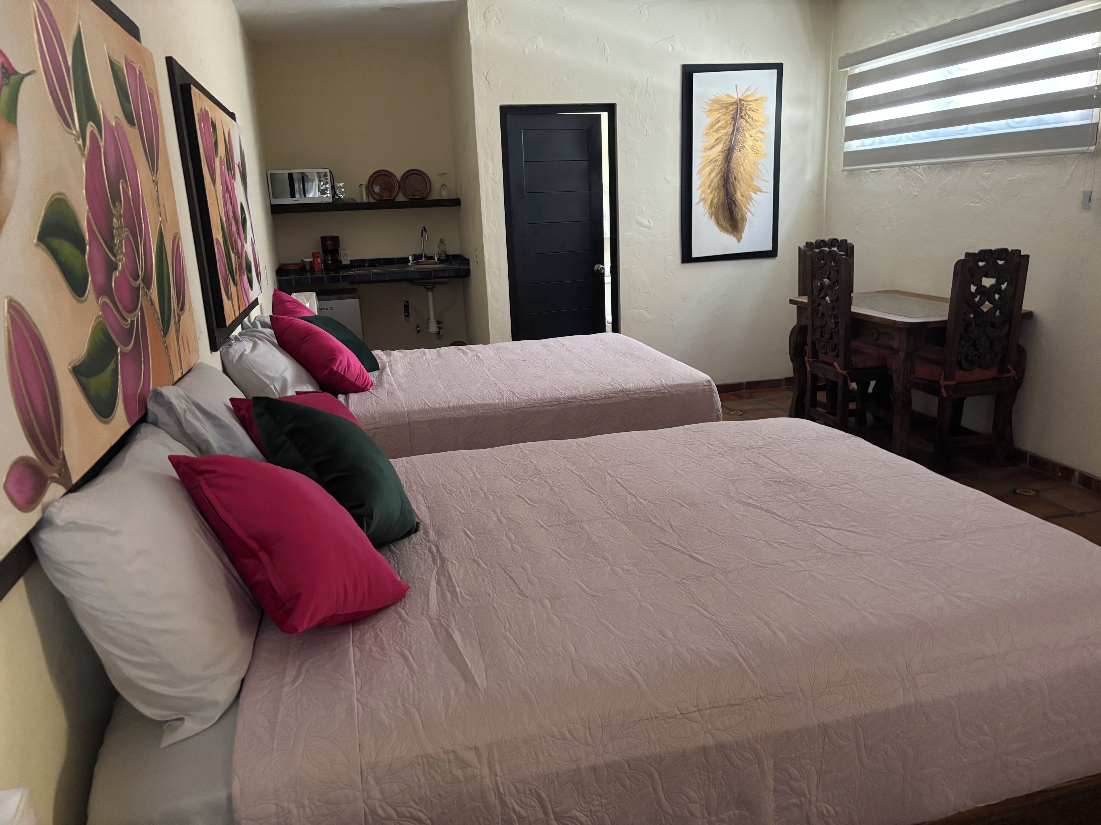 A room with two beds, a small kitchenette, a dining area with a table and chairs, and artwork on the walls.