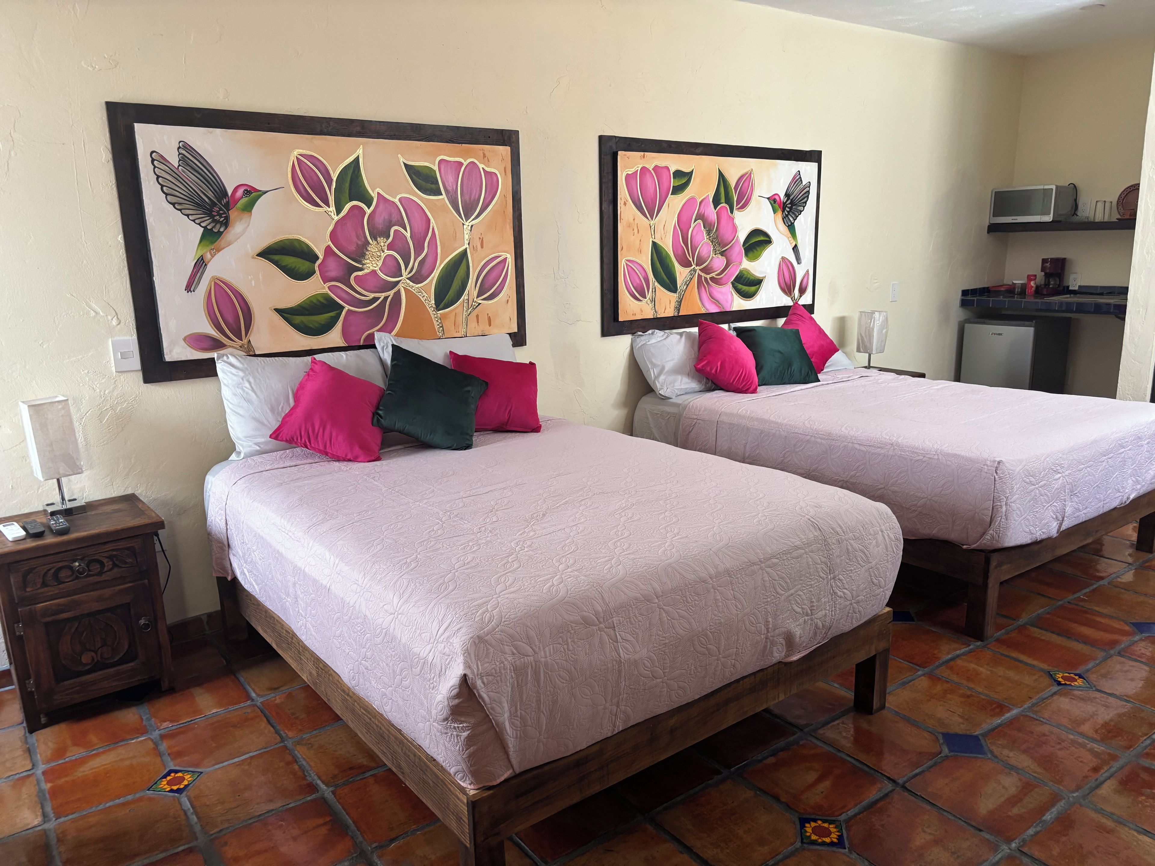 Habitación de hotel con dos camas, cada una con cubrecama y almohadas rosas, una mesita de noche con lámpara, y una pared decorada con dos cuadros de motivos florales.