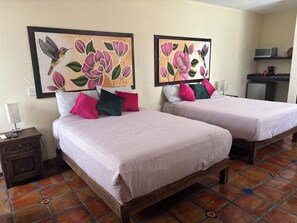 A hotel room with two beds, each with a pink bedspread and pillows, a nightstand with a lamp, and a wall with two framed floral paintings.