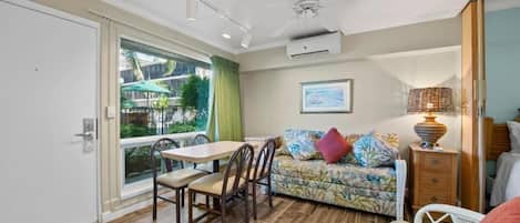 A room with a dining table, chairs, a sofa with cushions, a lamp, and a ceiling fan.