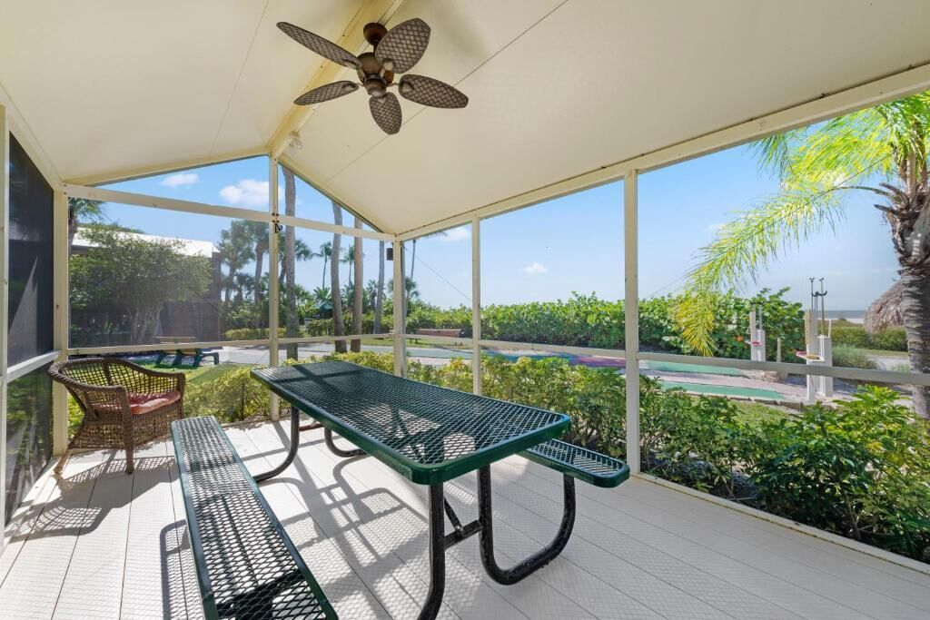 Un espace extérieur abrité, équipé d’une table et de chaises, d’un ventilateur de plafond, et offrant une vue sur une route et la verdure environnante.