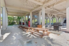 Uma área de piquenique coberta ao ar livre com mesas e bancos de madeira, churrasqueira e cooler Coca-Cola.