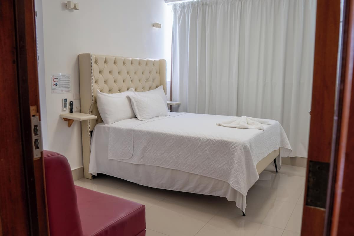 A neatly made bed with a white bedspread and pillows, a red chair, and a bedside table with a lamp.