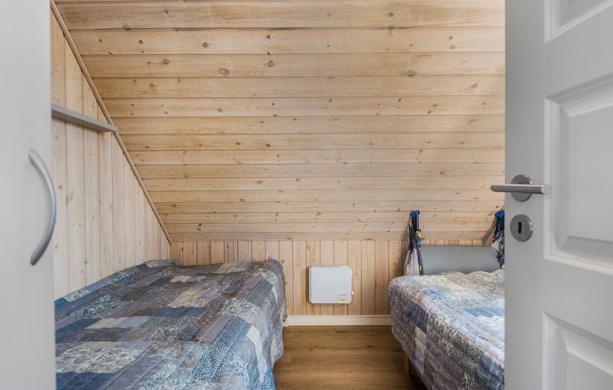 Una stanza accogliente e intima, con pareti in legno e soffitto spiovente. Sono presenti due letti con coperte a motivi decorativi, un termosifone bianco e una porta con maniglia.