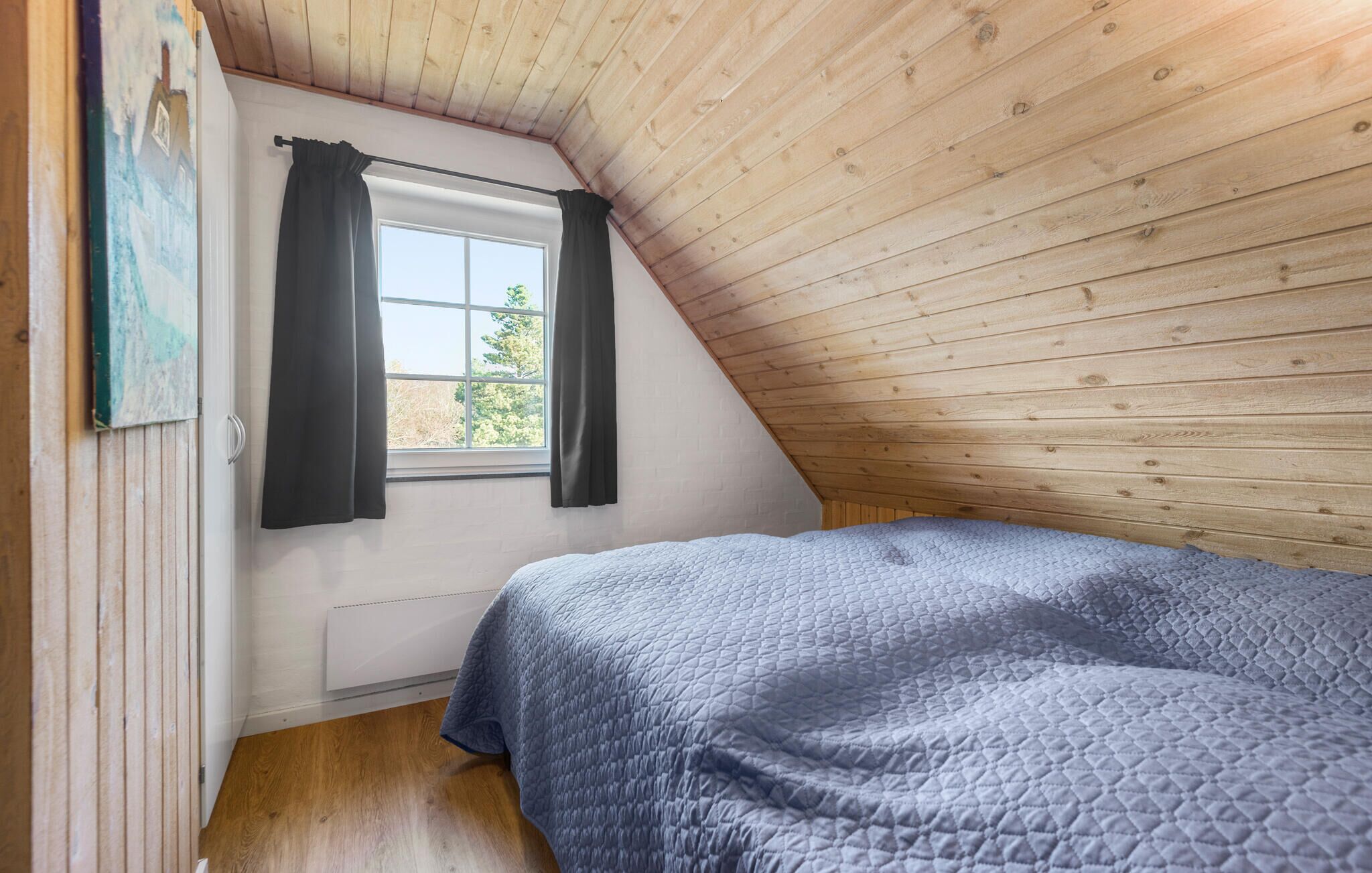 A bedroom with a sloped ceiling, a window with curtains, a bed with a quilted blanket, and wooden walls.