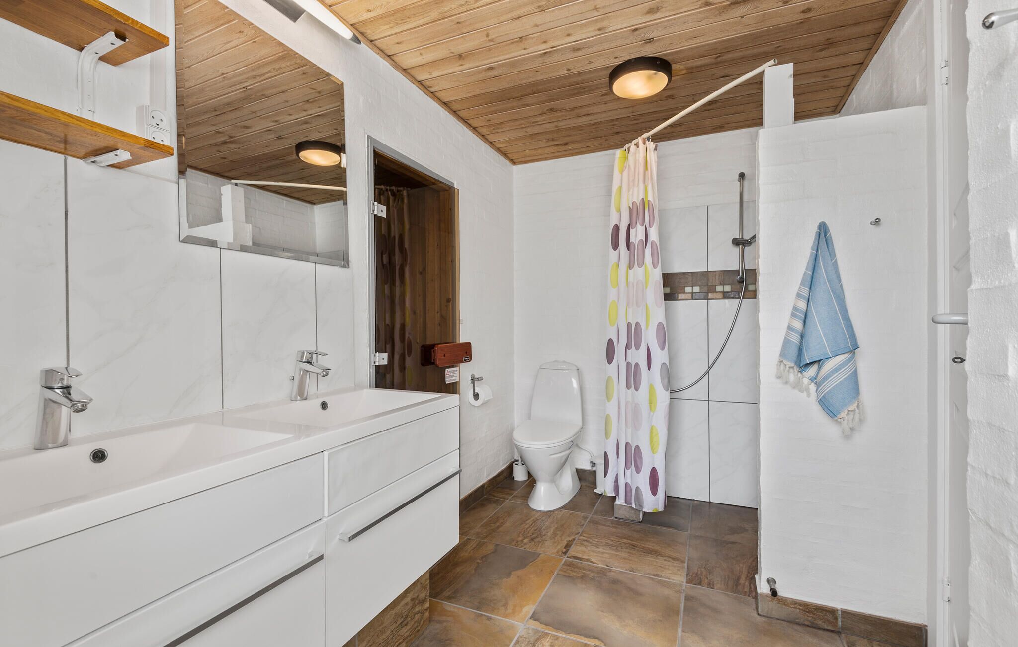 Een moderne badkamer met een houten plafond, een grote spiegel, een witte wastafel, een toilet en een douche met een stippen gordijn.
