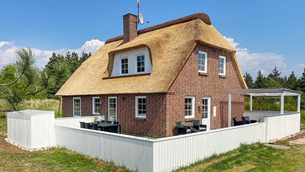 Ein Haus mit einem Strohdach, Backsteinmauern und einem umzäunten Garten mit Sitzgelegenheit.