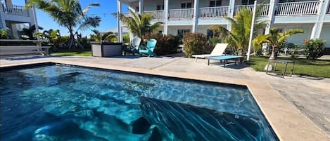 A swimming pool with clear blue water, surrounded by a poolside area with lounge chairs and a building with balconies in the background.