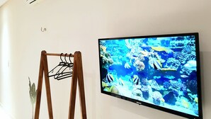 A television mounted on a wall displaying an underwater scene, a wooden easel with a plant, and a ceiling vent. - Estudio Mar'Bella 2 min Praia Caminho de Moisés com Garagem e ArCond (Maragogi)