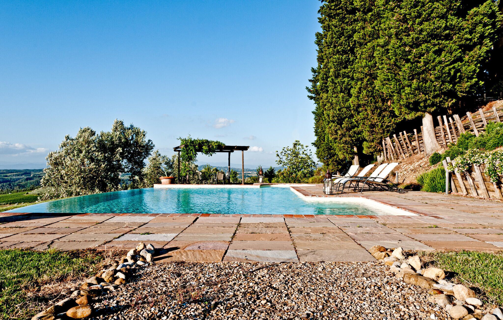 En swimmingpool med en pergola och solstolar, omgiven av ett stenbelagt däck och med utsikt över landskapet.