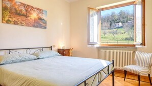 A bedroom with a bed, a chair, a window with a view of a house and trees, and a painting on the wall. - 2 bedroom cozy home in Varese Ligure (Varese Ligure)