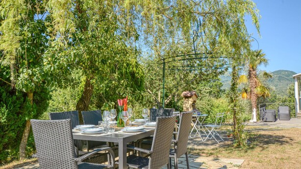 An outdoor dining area with wicker chairs and tables set up under a tree, surrounded by lush greenery and a mountain view. - 2 bedroom cozy home in Varese Ligure (Varese Ligure)