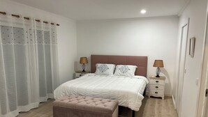 A bedroom with a bed, two bedside tables, a bench, and a window with sheer curtains.