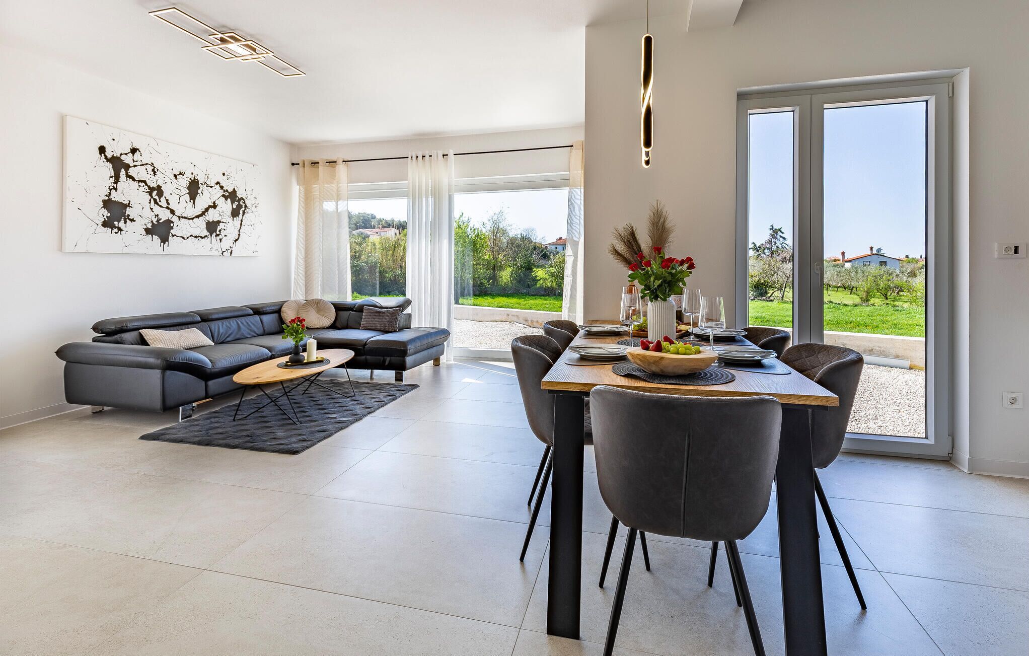 Ein modernes Wohnzimmer mit einem grauen Sofa, einem Esstisch für vier Personen und einem großen Fenster mit Blick auf Grünflächen und Gebäude.