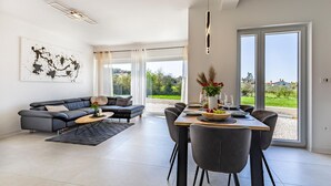 A modern living room with a grey sofa, a dining table set for four, and a large window offering a view of greenery and buildings. - 2 bedroom gorgeous apartment in Pula (Pula)