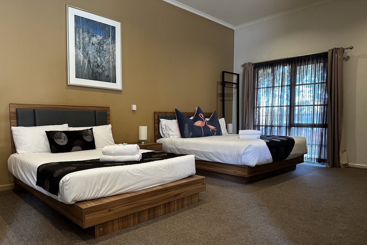 A hotel room with two beds, a large window with curtains, a painting on the wall, and a carpeted floor.