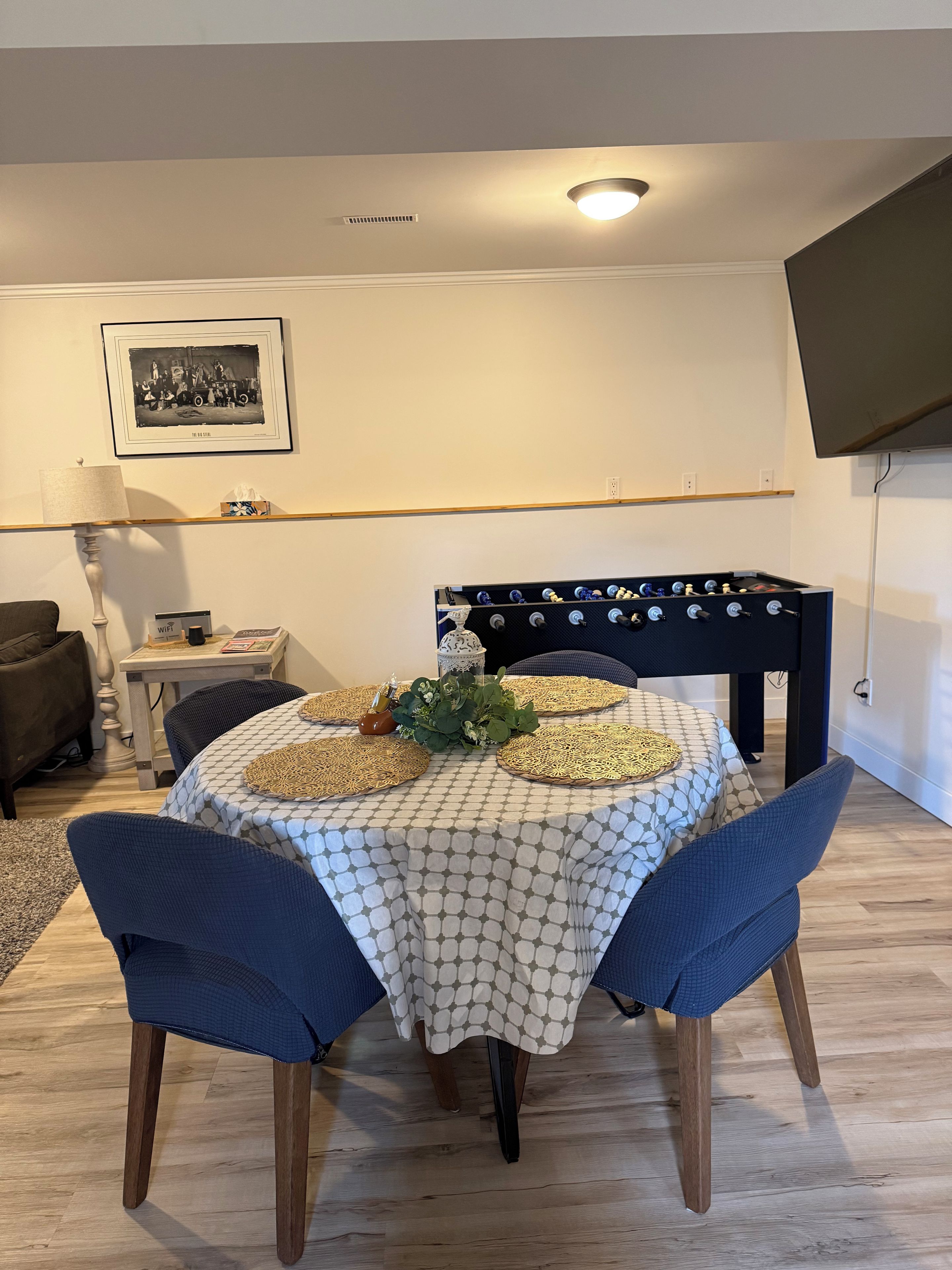 Un comedor con una mesa dispuesta para cuatro, un sofá, un televisor montado en la pared y un cuadro enmarcado sobre la mesa.