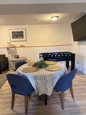 Un comedor con una mesa dispuesta para cuatro, un sofá, un televisor montado en la pared y un cuadro enmarcado sobre la mesa.