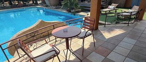 Un espace aménagé au bord de la piscine, doté d’un sol carrelé, d’une table ronde, de deux chaises et d’un coin salon surmonté d’une pergola.