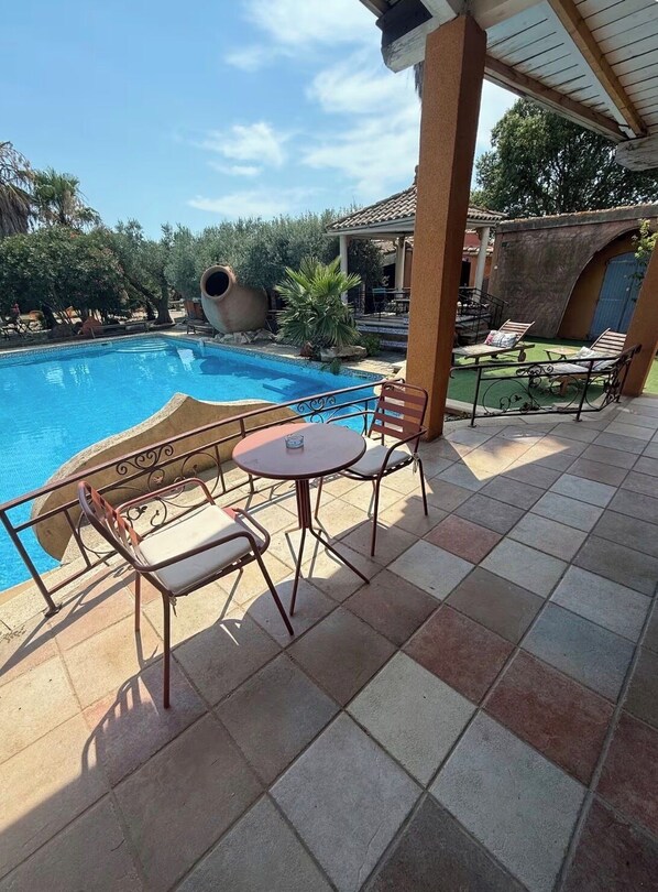 A poolside area with a tiled floor, a round table, two chairs, and a lounge area with a pergola overhead.