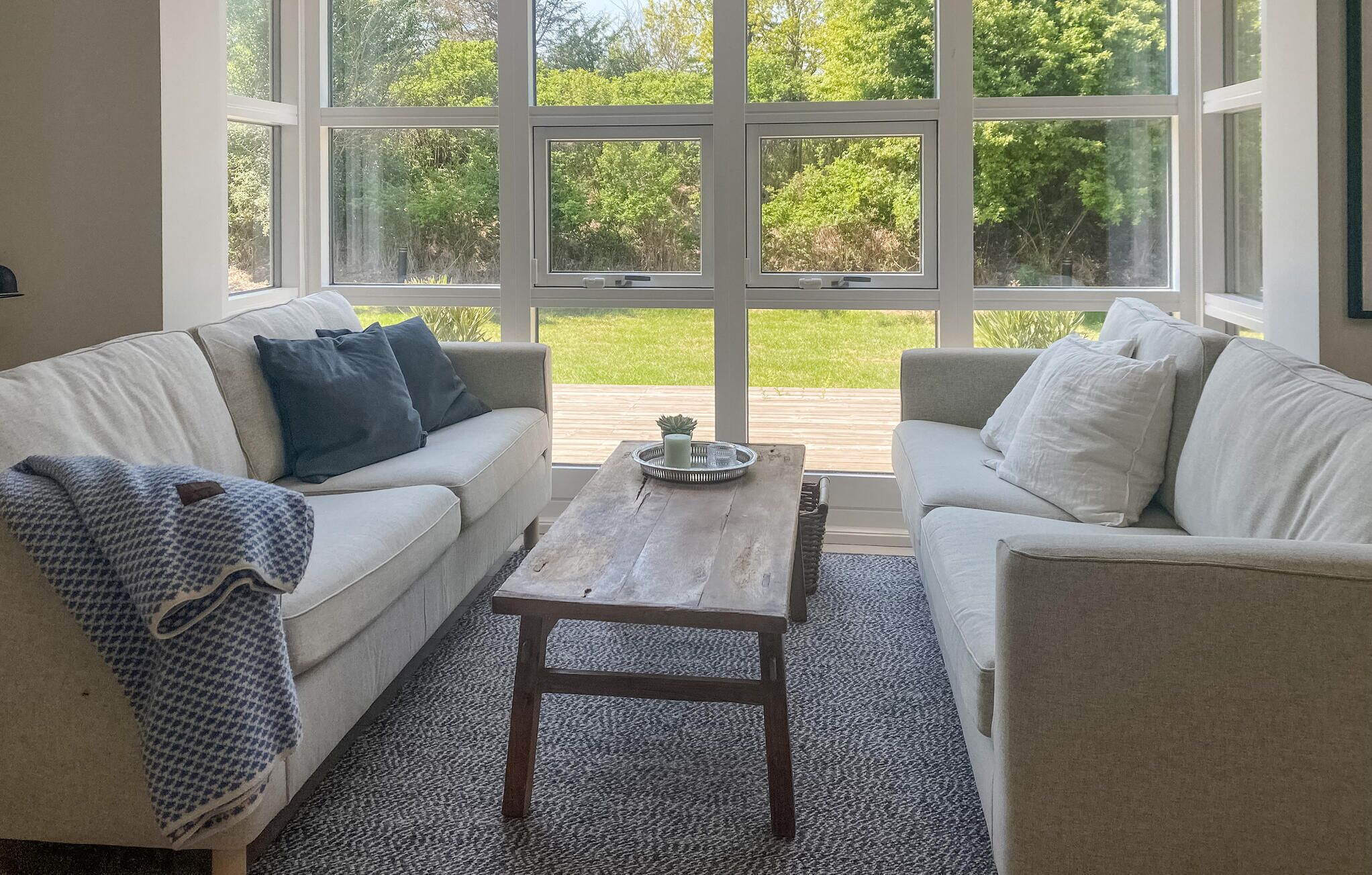Une pièce avec deux canapés blancs, une table basse en bois et de grandes fenêtres donnant sur un paysage verdoyant.