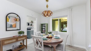 A dining area with a wooden table set for four, surrounded by white chairs. A wooden console table with a mirror and decorative items is to the left. A modern pendant light hangs above the table.