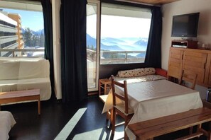 A bedroom with a bed, a wooden bench, a small table, a TV, and a view of mountains through a large window. - Family Escape in the Alps (Chamrousse)