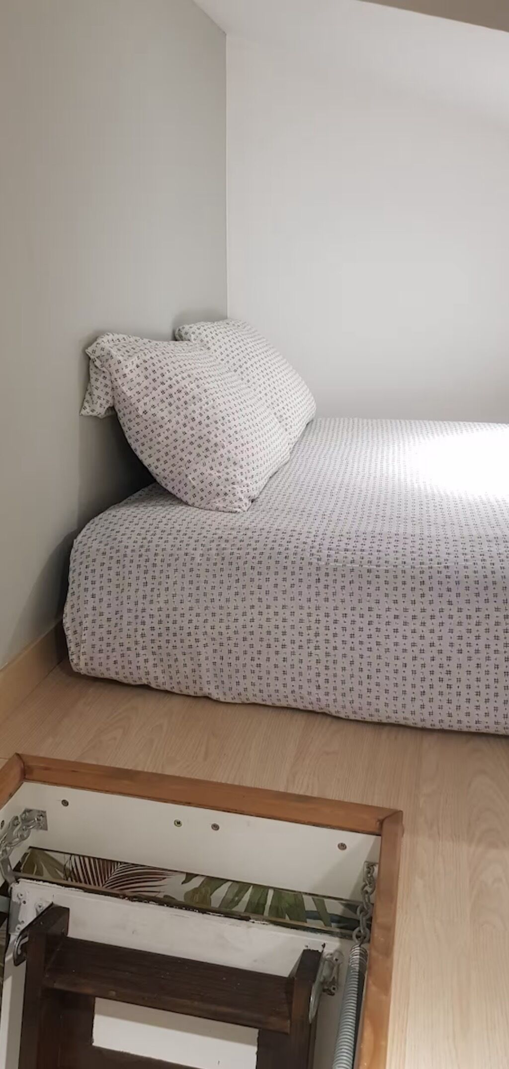 Un petit lit bien fait, avec une couverture à motifs et un oreiller, placé contre un mur uni dans une pièce au plancher en bois.