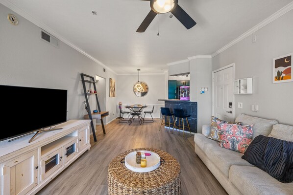 A living room with a flat-screen TV, a woven ottoman, a ceiling fan, and a kitchen in the background.