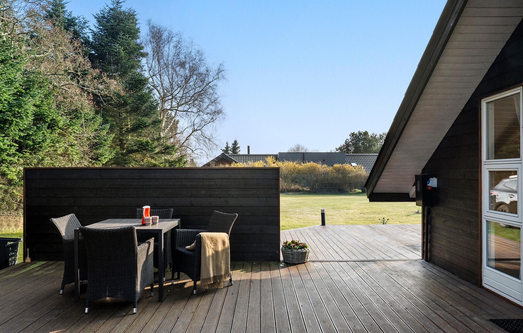 Een terras met een tafel en stoelen, een houten terras en uitzicht op een huis en bomen.