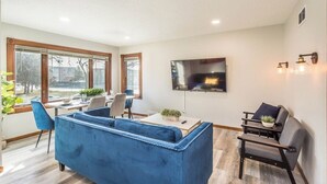 Un saló modern amb un sofà blau, una zona de menjador amb taula i cadires, un televisor penjat a la paret i una finestra gran que deixa entrar llum natural.