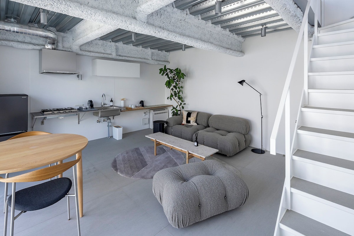 Une cuisine moderne équipée d’une cuisinière intégrée, d’un évier et d’un petit réfrigérateur. On y trouve un coin repas avec une table et des chaises en bois, ainsi qu’un canapé gris avec une plante à côté. Un escalier est visible sur le côté droit.