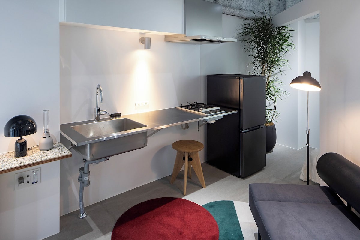 Une cuisine compacte avec un évier en acier inoxydable, un réfrigérateur noir et un petit tabouret.
